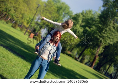 daughter girl sitting on her mother's shoulders in summer park pretended to be an airplane
