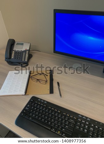 Desk with telephone, keyboard, computer monitor, notebook, pen and eyeglasses.