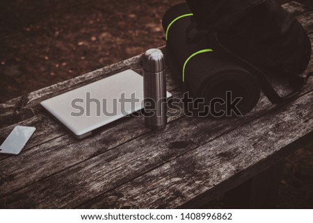 Silver laptop, smartphone , black backpack, mat and thermos on the wooden table in the forest. Sunset at background.