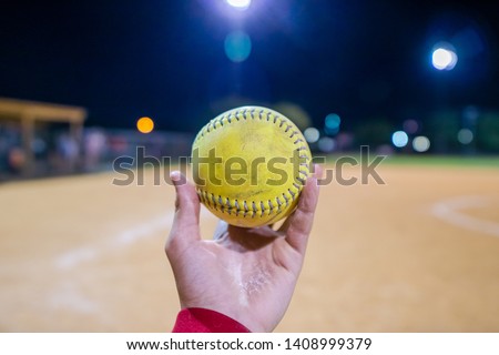 A person holding a softball in front of a softball field at night. 