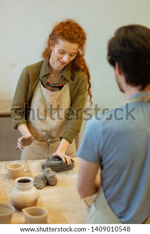 Creating pot. Smiling long-haired student in green shirt pressing piece of clay while learning new techniques in pottery