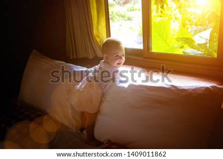 A laughing little girl in a white dress is standing on a bed against the background of the window behind which the jungle and sunny. A happy child of eight months leaned on the window sill.