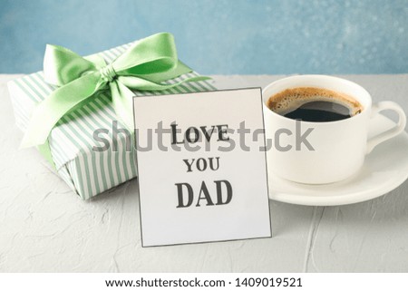 Cup of coffee, gift box with green ribbon and inscription Love you DAD on white table against blue background, space for text