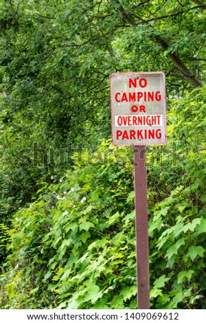 No camping or overnight parking sign with red lettering on rural road beside green trees.