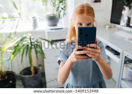 Teenage redhead girl in a blue dress is taking picture with her smartphone in a workshop.