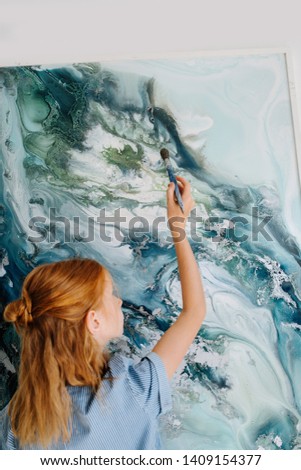 Teenage redhead girl in blue dress is pretending to paint on large canvas on the wall in a workshop. Like she's making little final brush strokes to finish perfect painting.