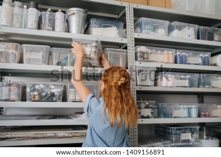 Teenage freckled redhead girl in a blue dress is carrying plastic box with little dry paint jars. She is putting it on the highest tier of a metal rack. Royalty-Free Stock Photo #1409156891