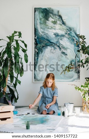 Teenage redhead girl in blue dress is making quality painting on large canvas on floor in a workshop. She's using paint pipette to drip water on painting. Beautiful art work in background.