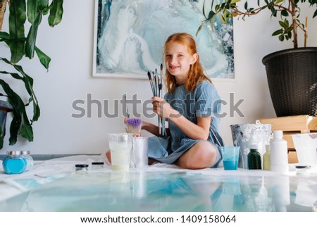 Teenage redhead girl in blue dress is posing, smiling, playing with brushes in a workshop. She is sitting on floor behind canvas with plastic paint jars around it.