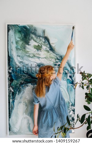 Teenage redhead girl in blue dress is pretending to paint on large canvas on the wall in a workshop. Like she's making little final brush strokes to finish perfect painting. Reaching high.