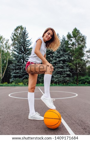 Beautiful young blonde girl dressed in white t-shirt, shorts and sneakers, plays with ball on a basketball court outdoors.