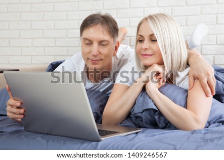 Smiling attractive couple lying on bed using laptop communicating online at home, happy woman and man typing on computer, enjoying internet shopping or chatting in social networks in modern bedroom