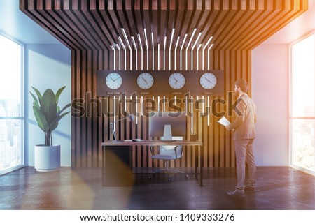 Young businessman with documents standing in modern office with wooden and white walls, computer table and clocks showing world time. Toned image