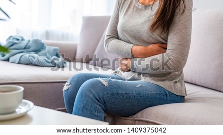 Young woman in pain lying on couch at home, casual style indoor shoot. Young woman with stomach ache sitting on bed. Young woman with stomach pain Royalty-Free Stock Photo #1409375042