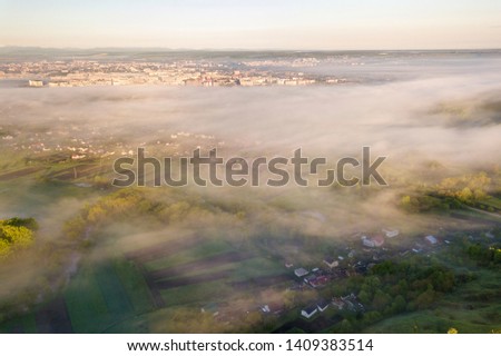 View from above of white fog on village house roofs among green trees under bright blue sky. Spring misty landscape panorama at dawn.