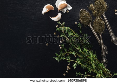 top view of dried thyme in silver spoons near green herb, white pepper and garlic cloves on black background with copy space