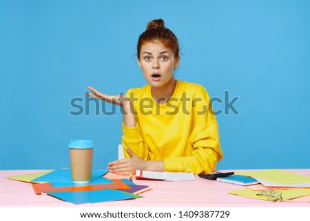 Surprised woman shows her hand to the side and sits at a pink table on a blue background, a folder with documents designer manager
