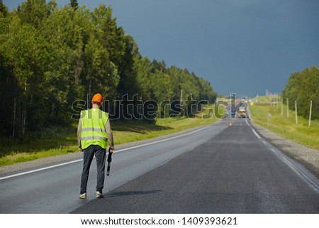The road worker with a rod regulates the movement of the transport in the repaired part of the roadway. Riding cars on the background of the rain sky, dangerous, can knock down
