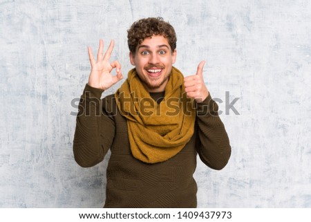Blonde man over grunge wall showing ok sign and thumb up gesture