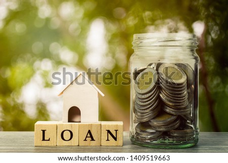 Saving money, home loan, mortgage, a property investment for future concept. A small residence house on wooden block and money coins in jar on wood table and space with green background and light.