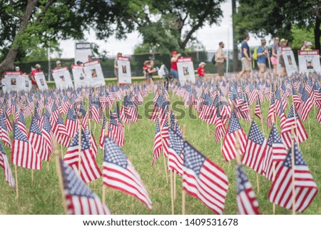Close-up view of lawn American flag with row of people from Memorial Day March event in Dallas, Texas, USA. Blurry crowded family members carry fallen heroes banners pictures placards in parade