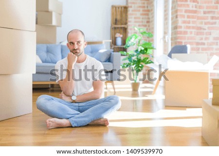 Young bald man sitting on the floor around cardboard boxes moving to a new home with hand on chin thinking about question, pensive expression. Smiling with thoughtful face. Doubt concept.