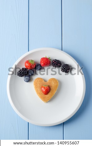 Overhead shot of a heart shaped breakfast pancake underneath an arc of Summer fruits and topped with a cut strawberry.  Food set on a white china plate with a painted wood planked table underneath.