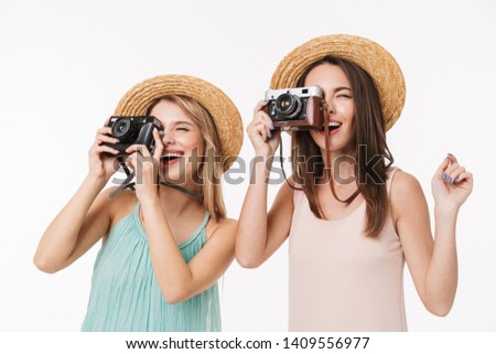 Two cheerful pretty young girls standing isolated over white background, wearing straw hats, taking pictures with photo cameras