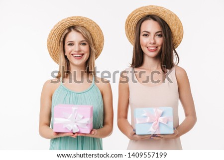 Two cheerful pretty young girls wearing summer outfit standing isolated over white background, showing present boxes