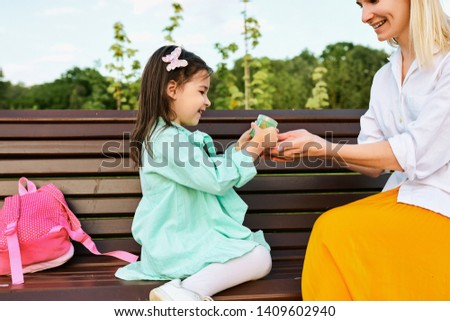 Outdoor image of happy cute little girl got a gift from her mother sitting on the bench in the park. Happy mothers day! Beautiful young woman smiling and playing with her child. Motherhood, childhood