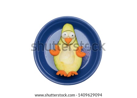 Food art creative concept. Funny penguin made of avocado, cucumber and carrots on a blue plate over white background.  Creative healthy food concept, an avocado cut in half with  funny googly eyes.