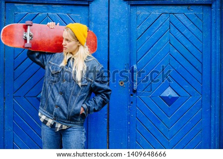 Blond hipster woman in yellow hat and blue jeans jacket holding longboard on her shoulder over old blue woden wall background, outdoor. Girl looks aside. Copy space text. Lifestyle concept