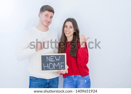Beautiful young couple over white background holding blackboard with new home text pointing and showing with thumb up to the side with happy face smiling