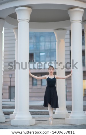 Ballerina in a black dress is a beautiful pose. A beautiful young woman in a black bodysuit is dancing in an elegant classical dance on a background of columns.