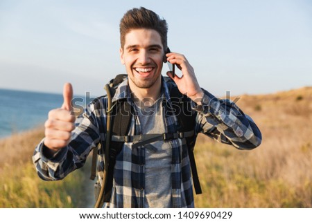 Photo of handsome happy young man outside in free alternative vacation camping talking by mobile phone showing thumbs up gesture.