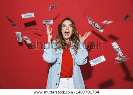 Portrait of a cheerful stylish young woman wearing denim jacket standing isolated over red background, standing under money banknotes rain