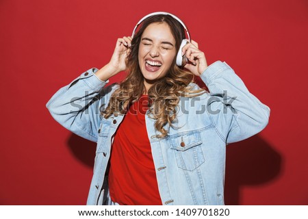 Portrait of a cheerful stylish young woman wearing denim jacket standing isolated over red background, listening to music with headphones, dancing Royalty-Free Stock Photo #1409701820