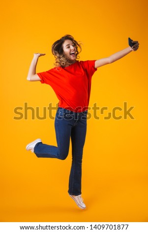 Full length portrait of a lovely cheerful playful young woman jumping isolated over yellow background, posing, taking a selfie