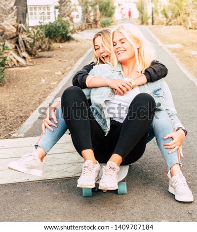 Happy young people couple of friends girls have fun together riding a skateboard on the road for outdoor leisure funny activity - friendship and craziness lifestyle concept for alternative millennial