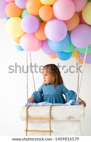 a little girl is standing in the basket of a balloon. the baby is dressed in a denim dress. happy childhood. toy balloon made of colored balls. photo zone to celebrate a birthday