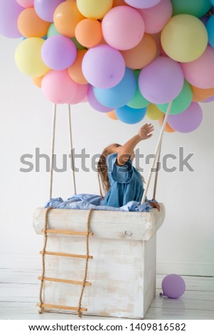 a little girl is standing in the basket of a balloon. the baby is dressed in a denim dress. happy childhood. toy balloon made of colored balls. photo zone to celebrate a birthday