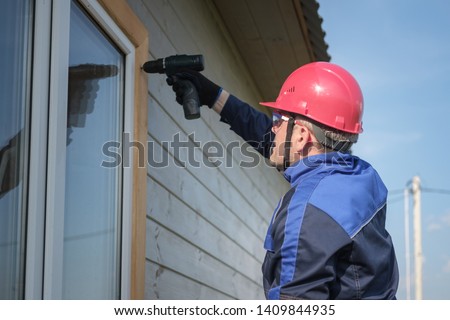 European male builder in overalls assembles a window casing of a wooden house with a screwdriver. construction and repair of wooden frame houses Royalty-Free Stock Photo #1409844935