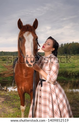 A woman in a retro dress is standing next to a horse. The girl and the horse in the photo. The girl in retro style holds a horse by the bridle.