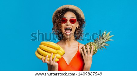 Tropical summer fruits. Emotional black girl holding bananas and pineapple, blue studio background