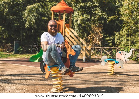 Playful crazy man (dad) riding wooden rocking horse in a park - adult guy (hipster with beard) having fun on a playground Royalty-Free Stock Photo #1409893319
