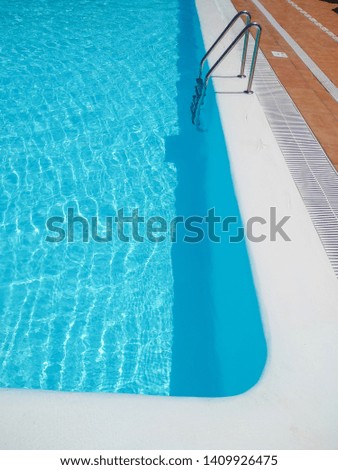 corner of a swimming pool of a touristic place full of clear and transparent water. The edge of the pool is in white color and there are some brown tiles around and a metallic ladder. Vertical photo