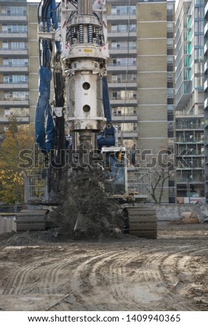 Large machine digs a round hole on a construction site.  Autumn day and fall colors with trees having yellow leaves in background.