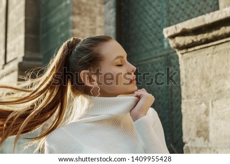 Fashionable woman with long hair in ponytail hairstyle wearing trendy hoop earrings, white turtleneck sweater, posing in street of European city. Copy, empty space for text Royalty-Free Stock Photo #1409955428