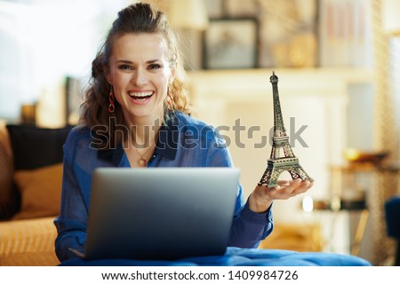 smiling stylish woman with a souvenir of the eiffel tower with laptop in the modern house in sunny summer day. A fake website is made for illustrative purposes.