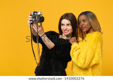 Two young women friends european and african american in black yellow clothes standing posing isolated on bright orange wall background, studio portrait. People lifestyle concept. Mock up copy space